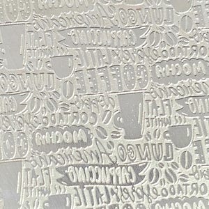 May include: A textured, off-white surface with an embossed pattern of coffee-related words and images. The design includes words like "coffee," "latte," and "cappuccino," along with coffee cup illustrations. The overall effect is a repeating, all-over design.