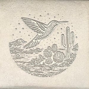 May include: A detailed, etched design of a hummingbird in flight over a desert landscape with cacti and mountains. The image is in shades of gray and white, with a circular composition and a starry sky.