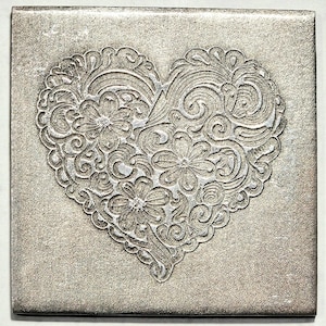 May include: Square, textured, silver-colored tile with a raised heart design. The heart is filled with floral and swirl patterns. The tile has a slightly distressed appearance, adding to its vintage charm.