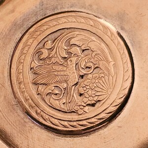 May include: A close-up of a circular, copper-colored plate with an intricate design of a bird and floral motifs. The design is engraved into the surface of the plate.