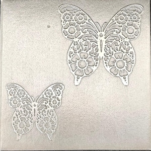 May include: A square, textured, silver-colored surface features two detailed butterfly designs. The butterflies are white, with intricate floral and swirling patterns within their wings. The larger butterfly is centered, while a smaller one is in the lower left corner.