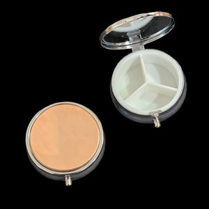 May include: A round, silver pill box with a mirrored lid. The box is open, revealing three compartments inside. The box is on a black background.