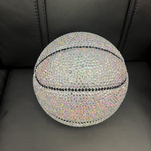 May include: A basketball covered in iridescent rhinestones. The ball has black lines and is set against a dark background. The rhinestones reflect a rainbow of colors, creating a sparkling effect.