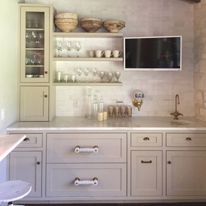 Puede incluir: Una barra de cocina blanca con encimera y armarios grises. Los armarios tienen pomos de cerámica blanca. La barra tiene un televisor integrado, estantes y un armario con puerta de cristal. Los estantes están llenos de vasos, cuencos y otros artículos de cocina.