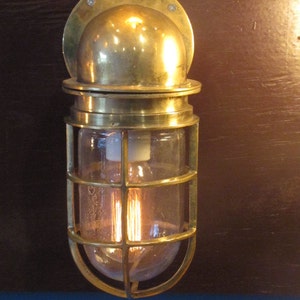 May include: A brass nautical bulkhead light fixture with a clear glass shade and a caged bulb. The light is mounted on a wall.