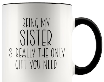 May include: White ceramic coffee mug with a black rim and handle. The mug has black text that reads "Being my sister is really the only gift you need."