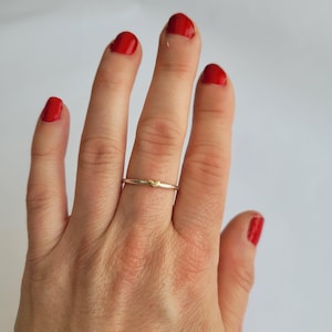 May include: A delicate gold ring featuring a small heart design. The ring is worn on the middle finger of a hand with red nail polish.