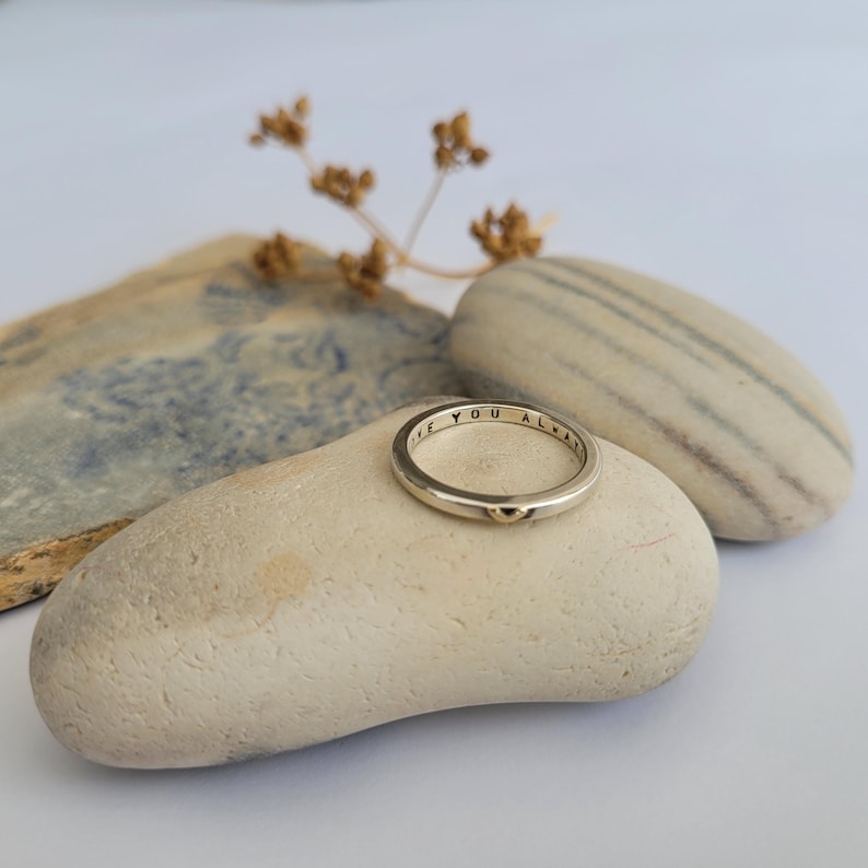 May include: A silver ring inscribed with the words "I love you always" and a small gold heart charm. The ring is resting on a light brown stone.