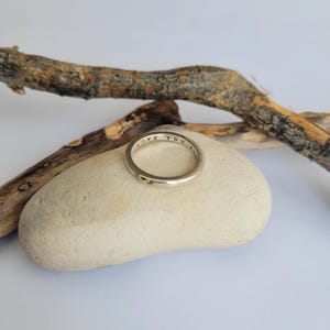May include: A silver ring with the words "LOVE YOU" engraved on the band. The ring is resting on a light brown rock.