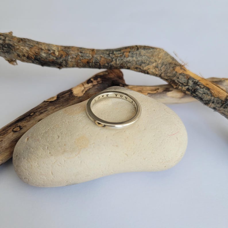 May include: A silver ring with the words "LOVE YOU" engraved on the band. The ring is resting on a light brown rock.