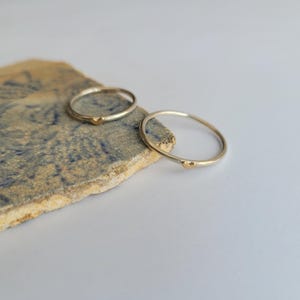 May include: Two thin silver rings with a small gold heart accent on each ring. The rings are on a textured, blue and brown surface.