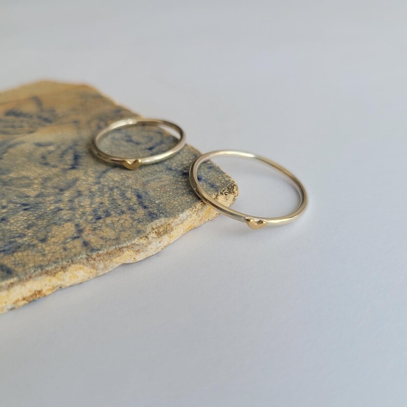 May include: Two thin silver rings with a small gold heart accent on each ring. The rings are on a textured, blue and brown surface.