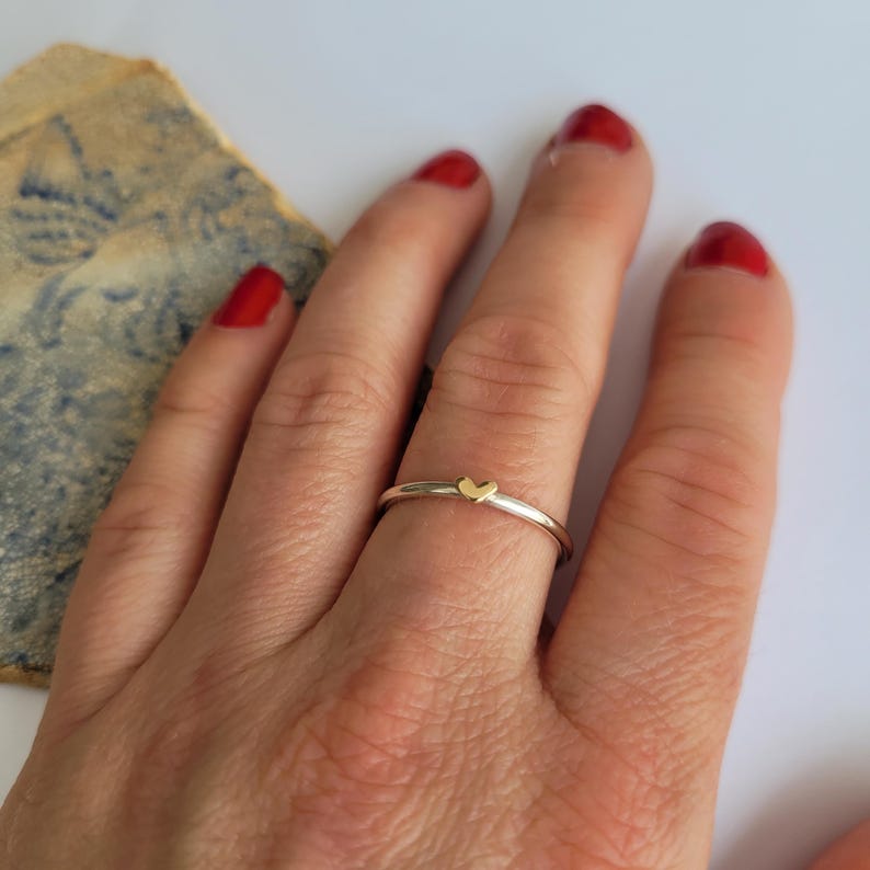 May include: A delicate silver band ring with a small gold heart charm.