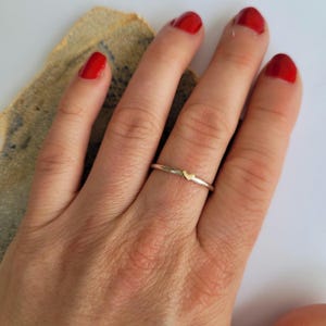 May include: A delicate silver ring with a small gold heart charm. The ring is worn on the finger of a hand with red nail polish.