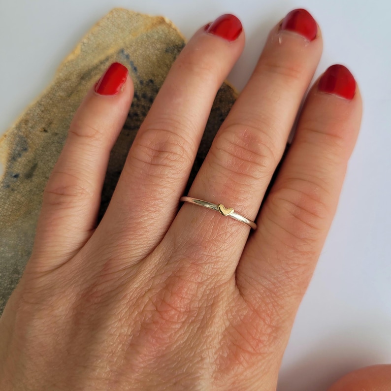 May include: A delicate silver ring with a small gold heart charm. The ring is worn on the finger of a hand with red nail polish.