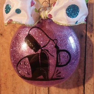 May include: Purple glitter Christmas ornament with a black silhouette of a pair of shoes. The ornament is decorated with a white ribbon with blue polka dots.