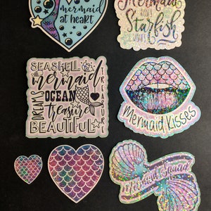 May include: Assortment of iridescent holographic mermaid themed stickers.  The stickers feature a variety of designs including a heart with a mermaid tail, a mermaid with the text "mermaid at heart", a mermaid with the text "Mermaid Kisses Starfish Wishes", a mermaid with the text "Seashell Mermaid Ocean Treasure Beautiful", a mermaid lip design with the text "Mermaid Kisses", and a mermaid with the text "Mermaid Squad".