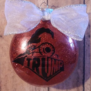 May include: Red glitter Christmas ornament with a black silhouette of a train engine and the word "TRUMP" printed on it. The ornament is decorated with a white sparkly bow.