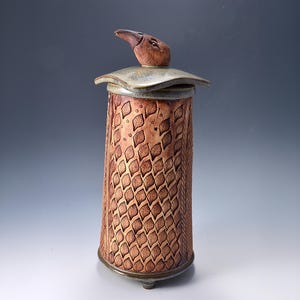 May include: A cylindrical ceramic jar with a textured surface featuring a repeating leaf pattern in shades of brown. The lid is topped with a sculpted brown bird figure and has a smooth, greyish-green finish with slightly upturned edges.