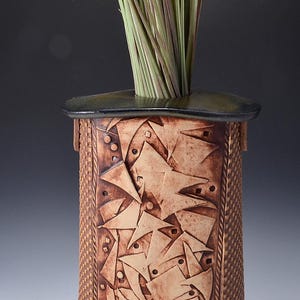 May include: A cylindrical ceramic vase with a textured, geometric design in shades of brown. The vase has a dark rim and base, and is filled with tall, green and purple grass. The vase is approximately 10 inches tall.