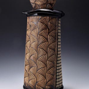 May include: A tall, decorative ceramic vessel with a black base and lid. The body features a repeating fan-like pattern in brown and black. A small, crescent-shaped handle sits atop the lid. The vessel has a rectangular section with a dotted pattern.