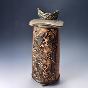 May include: A tall cylindrical ceramic vase with a flared rim and a textured surface. The body features intricate dark brown etched patterns of beetles and foliage against a lighter clay background. The base is supported by small feet.