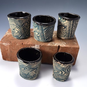 May include: Five ceramic cups with a dark blue and cream wave pattern and a smooth black interior. The exterior features a textured, carved design with repeating wave motifs and vertical braided lines, creating a distinct contrast between the dark blue recessed areas and the raised cream-colored surfaces.