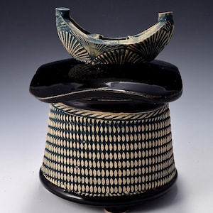 May include: A handcrafted ceramic sculpture with a unique design. The piece features a black base and a cylindrical body with a textured pattern. A black, curved element sits atop the body, supporting a crescent-shaped, blue-green accent with a fan-like design.
