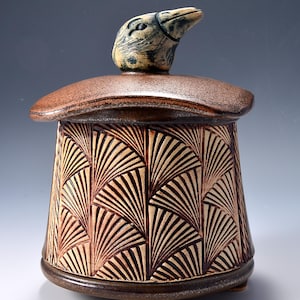 May include: A handcrafted ceramic jar with a lid and a bird-shaped handle. The jar features a fan-like pattern carved into the brown surface. The lid and base are a darker brown. The jar sits on three small feet.
