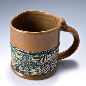 May include: A ceramic mug with a smooth, light brown glazed upper section and a textured lower section featuring a dark blue and cream-colored relief pattern of stylized ocean waves. The mug has a sturdy, curved handle and a cylindrical shape.
