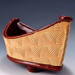 May include: A boat-shaped ceramic planter featuring a textured exterior with a repeating fan-like pattern in a light tan color. The rim, base, and interior are finished in a smooth, glossy dark reddish-brown glaze, supported by small feet.