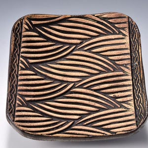 May include: A square, ceramic tray with a textured wave pattern in shades of brown and tan. The edges feature a braided design. This handcrafted piece is suitable for serving or display.
