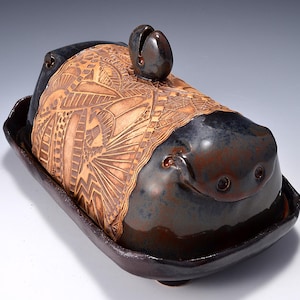 May include: A handcrafted ceramic butter dish with a lid. The dish is dark brown with a lighter brown, textured design on the lid. The lid has a small, rounded handle. The base is rectangular with rounded corners.
