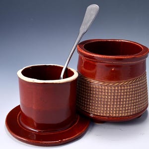 May include: A two-piece, dark red ceramic butter dish with a matching lid and a small silver spoon. The dish has a textured, woven pattern around the base. The lid is off, revealing the interior of the dish.