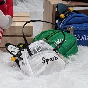 May include: A collection of six small, personalized backpacks in various colors, including pink, blue, green, red, and white. The backpacks have different embroidered names, including "Sparkle", "Howdy", "Snowflake", and "Buddy".