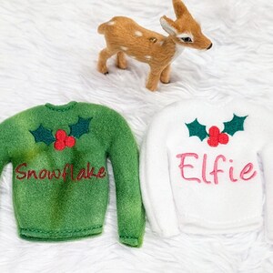 May include: Two green and white elf-sized Christmas sweaters with holly and berry embroidery. The green sweater says "Snowflake" and the white sweater says "Elfie".