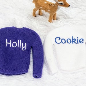 May include: Two elf-sized sweaters, one purple with "Holly" embroidered in white and one white with "Cookie" embroidered in blue.
