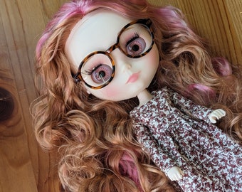 May include: A doll with long wavy hair featuring pink and light brown highlights. The doll has pale skin, pink lips, and large eyes with long eyelashes. It wears round tortoise-shell patterned glasses and a long-sleeved dress with a small floral print.