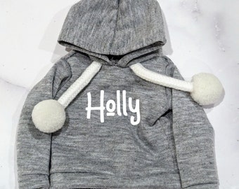 May include: A heather gray long-sleeve hooded sweatshirt featuring the name Holly printed in white lettering on the front. The hoodie includes thick white braided drawstrings that are finished with large, round, fuzzy white pom-poms at each end.