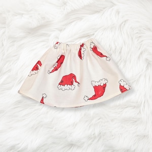 May include: A flared skirt with an elasticized waistband featuring an off-white base color and a repeating pattern of red Santa Claus hats with white trim. The fabric displays multiple illustrations of the hats oriented in various directions.