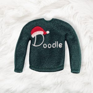 May include: A dark green long-sleeve sweater ornament featuring an embroidered red and white Santa hat positioned above the white stitched text that reads Doodle. The sweater has a round neckline and long sleeves with a textured fabric finish.
