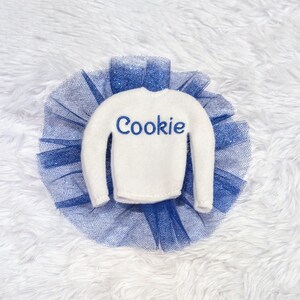 May include: A white elf-sized sweater with the word "Cookie" embroidered in blue on the front, paired with a blue tulle tutu.