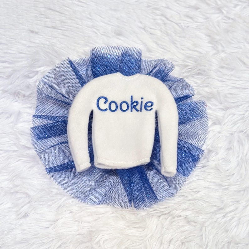 May include: A white elf-sized sweater with the word "Cookie" embroidered in blue on the front, paired with a blue tulle tutu.