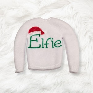 May include: A white long-sleeve sweater featuring an embroidered red and white Santa hat above the green stitched text Elfie. The garment has a round neckline and long sleeves, constructed from a soft, felt-like material with visible stitching.