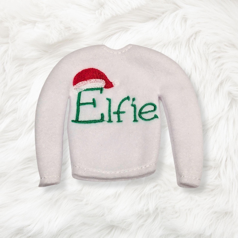 May include: A white long-sleeve sweater featuring an embroidered red and white Santa hat above the green stitched text Elfie. The garment has a round neckline and long sleeves, constructed from a soft, felt-like material with visible stitching.