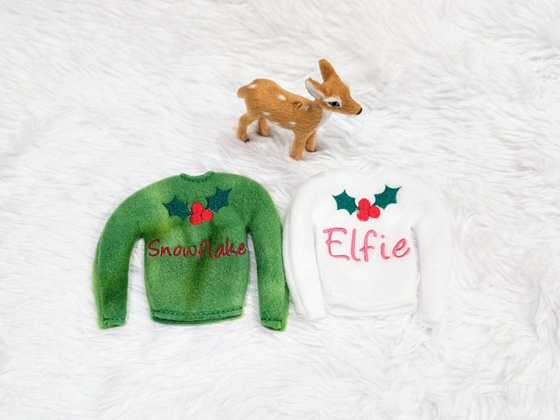 May include: Two green and white elf-sized Christmas sweaters with holly and berry embroidery. The green sweater says "Snowflake" and the white sweater says "Elfie".