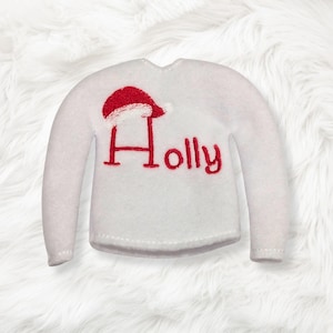 May include: A white long-sleeve sweater featuring the red embroidered text "Holly" on the front. The letter H is stylized with a red and white Santa hat resting on top of it. The sweater has a simple crew neckline and long sleeves.
