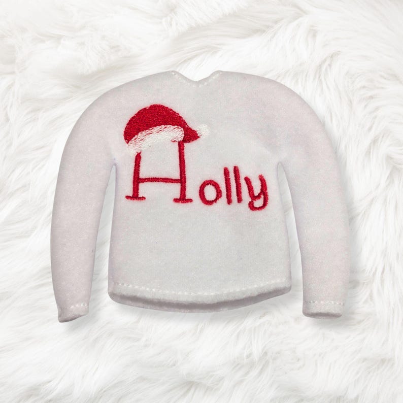 May include: A white long-sleeve sweater featuring the red embroidered text "Holly" on the front. The letter H is stylized with a red and white Santa hat resting on top of it. The sweater has a simple crew neckline and long sleeves.