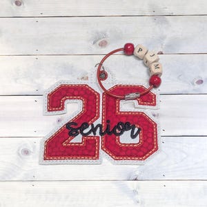 May include: A red and white embroidered patch with the number "25" and the word "Senior" in black script. A red beaded bracelet with wooden letter beads spelling out "PJM" is attached to the patch.