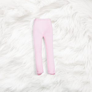 May include: A pair of light pink doll pants featuring a simple, fitted design with a high waist and straight legs. The garment is constructed from a solid, smooth fabric with visible stitching along the hem and inner seams of the pant legs.